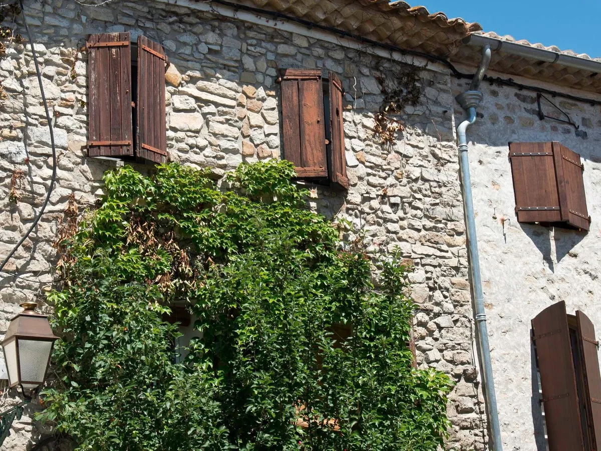 Façade ancienne en pierre de Provence avec volets battants en bois brun et vigne grimpante
