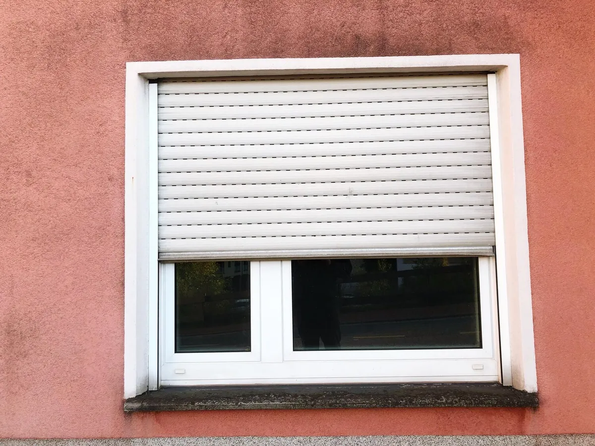 Fenêtre PVC blanche avec son volet roulant à lames arrêté à mi-hauteur sur une façade enduite rose